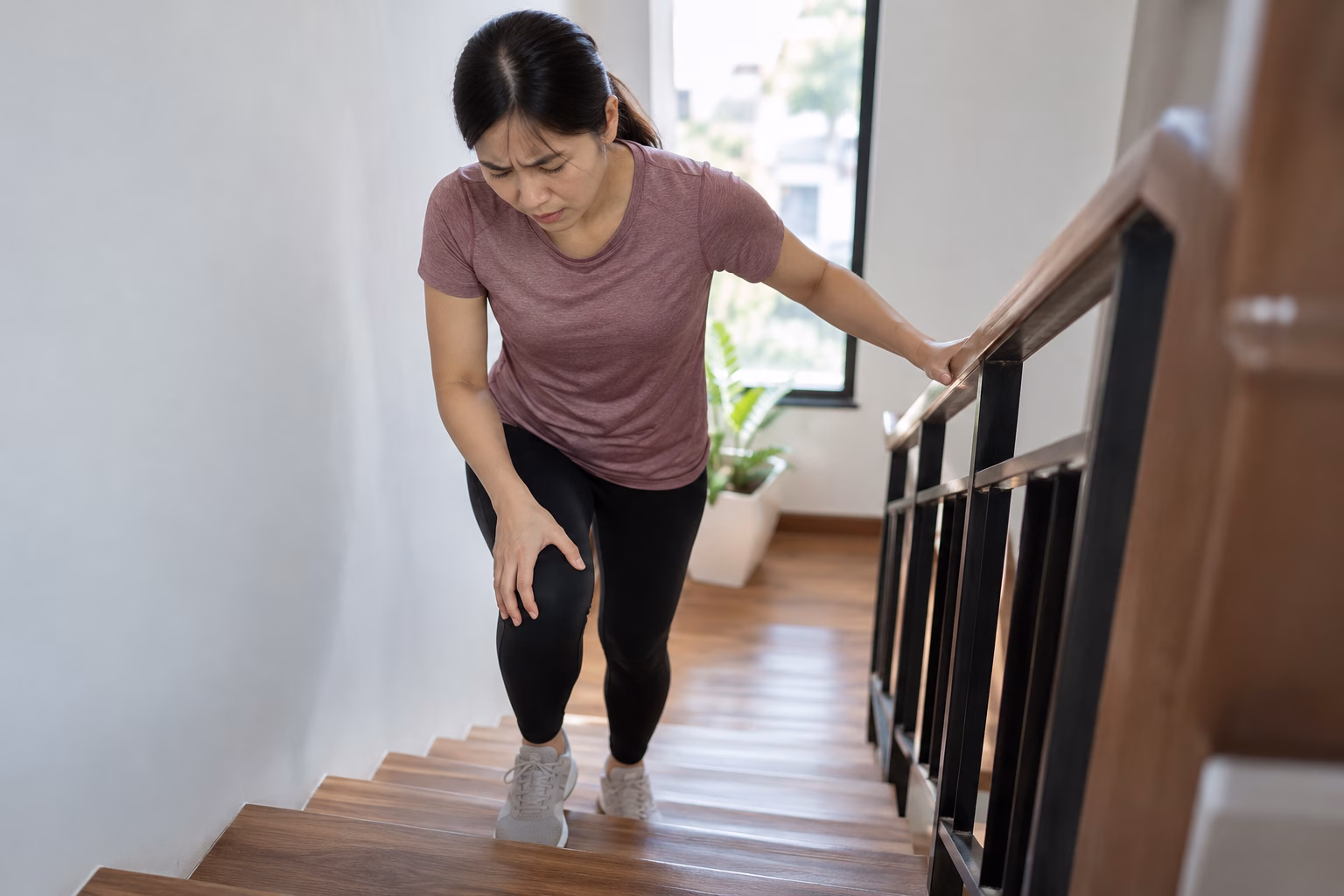 Woman experiencing knee pain while climbing stairs and using a handrail for support